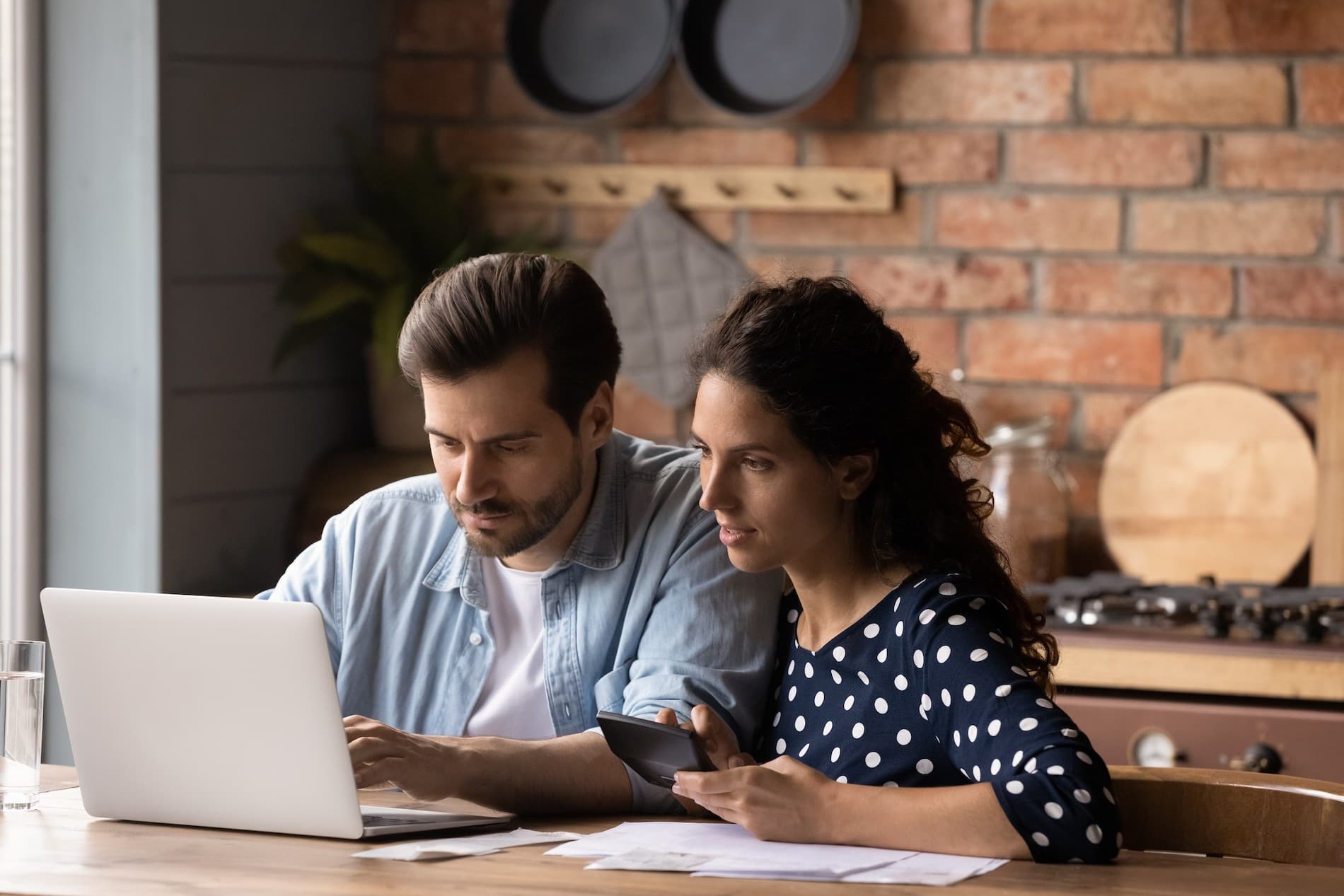 Two people at a kitchen counter working through paperwork on a laptop.